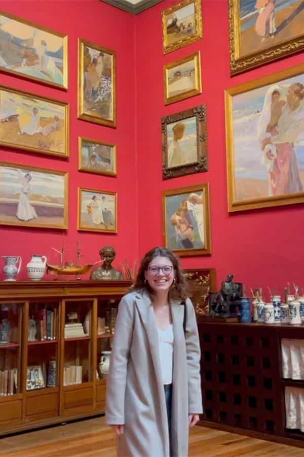 Martha Barnds, a female student smiles at the camera while standing in a room with red walls that are covered in framed paintings.